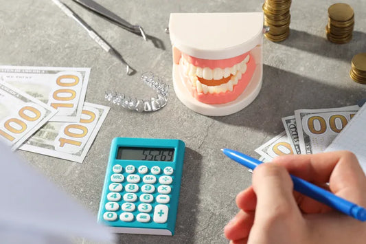 A hand with a pen, a calculator, a tooth model, aligners, and money on the table